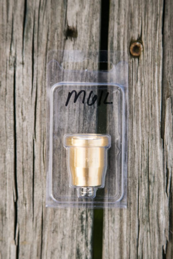 A brass-colored metal object is sealed in a clear plastic package labeled MUL in black marker. The package is placed on a weathered wooden surface with visible grain and cracks.