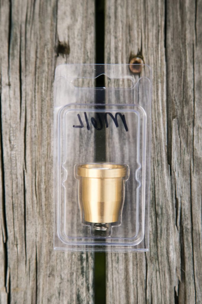 A small brass-colored metal part is packaged in a clear plastic case with a handwritten label that reads WOLF. The package rests on a weathered wooden surface with visible grain and cracks.