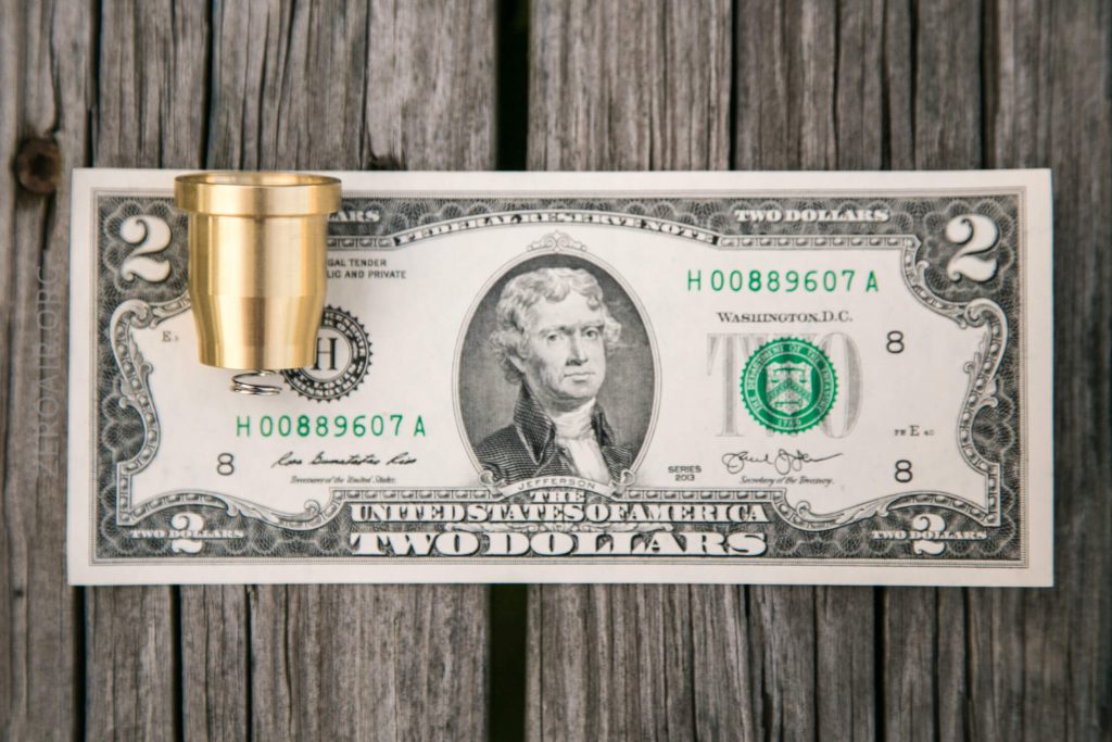 A two-dollar bill lies flat on a wooden surface with a small brass cup placed upside down on its left side.