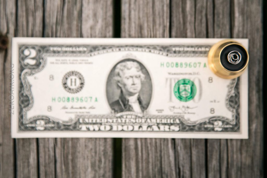 A two-dollar bill lies on a wooden surface with a gold and black vape device placed on its upper right corner.