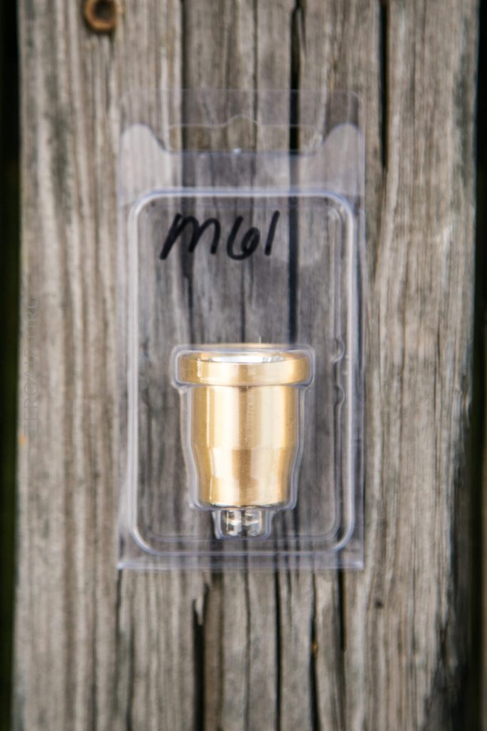 A small brass-colored metal part in a clear plastic package labeled “M61,” placed vertically against a weathered wooden surface.