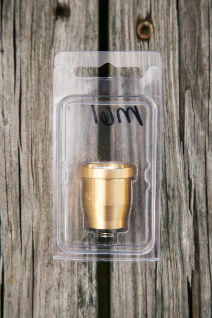 A small brass fitting in clear plastic packaging, labeled with 10M in black marker, placed on weathered wooden boards.
