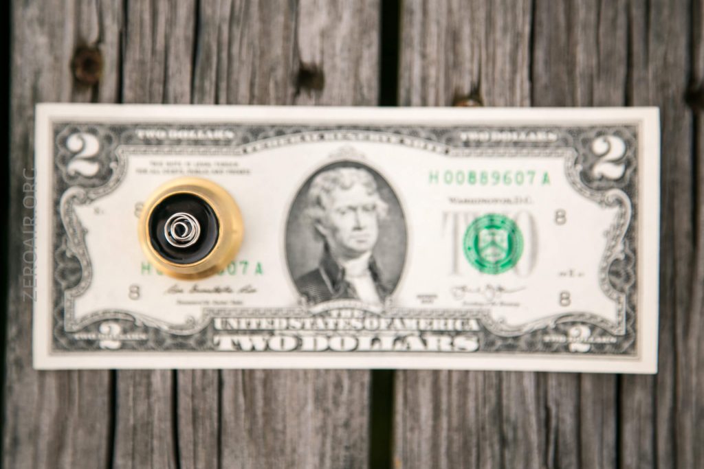 A US two-dollar bill with a small metallic object, possibly a bike valve or nozzle, placed vertically at the center, all resting on a weathered wooden surface.