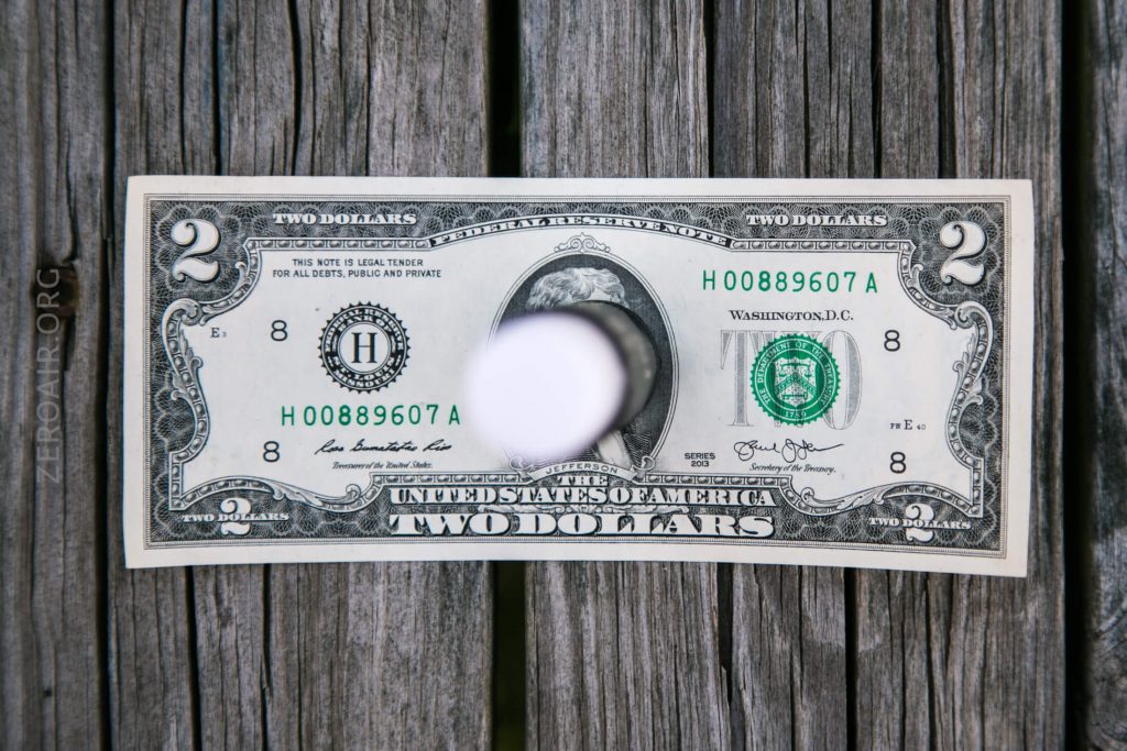 A two-dollar bill with a circular hole cut out of the center is placed on a wooden surface with visible grain.