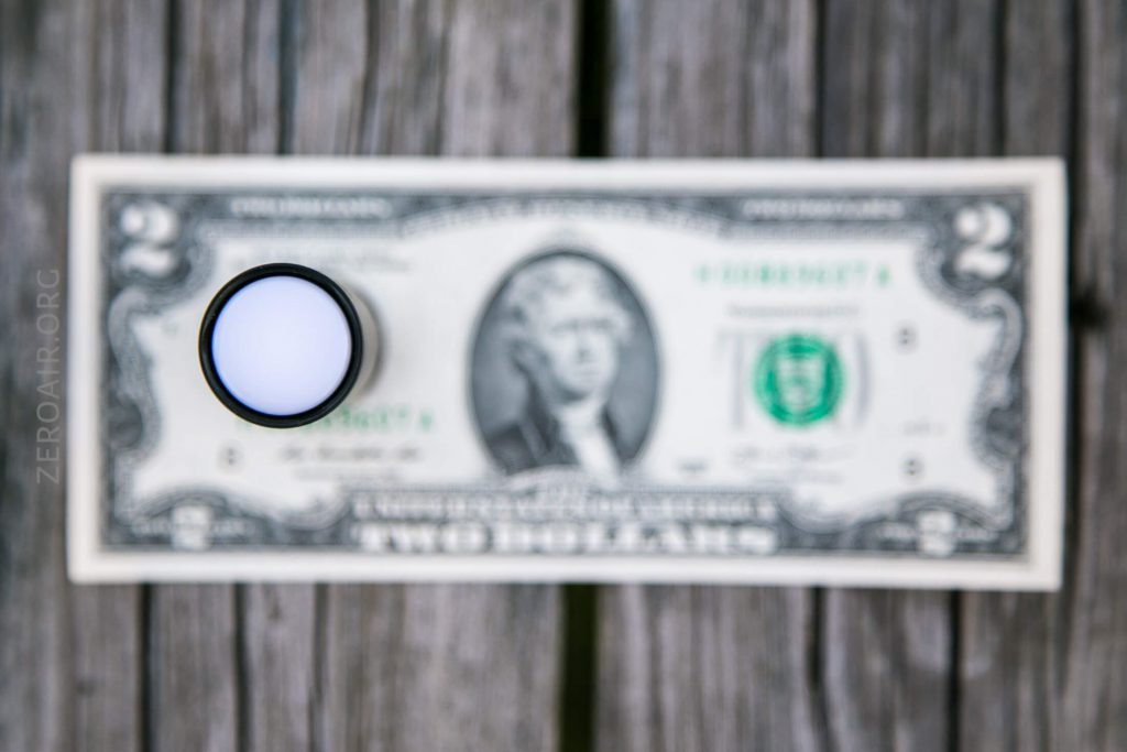 A round magnifying glass is placed over the portrait on a two-dollar bill, which is lying flat on a weathered wooden surface. The magnifier blurs and enlarges part of the bill underneath it.