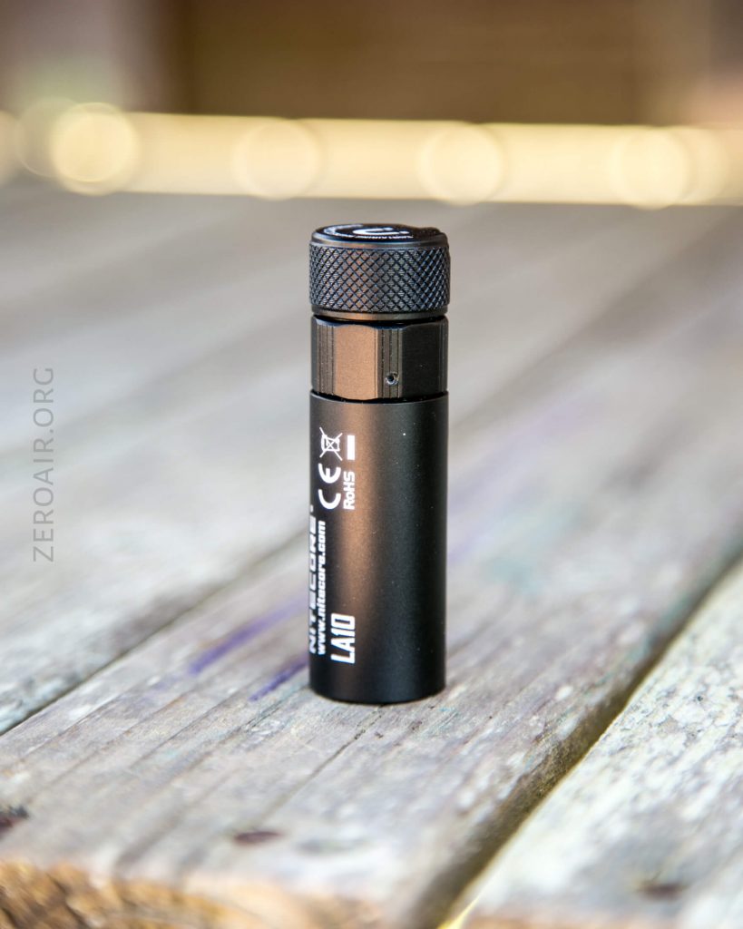 A small black flashlight labeled LAX10 stands upright on a weathered wooden surface, with ZEROAIR.ORG faintly visible on the left side of the image.