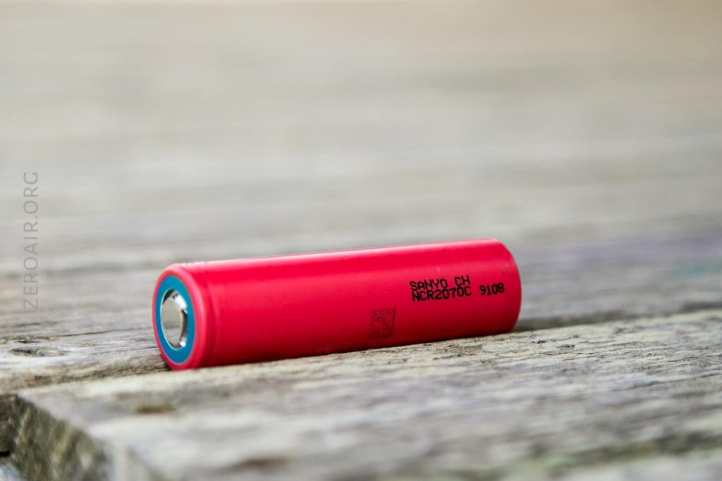 A red cylindrical battery labeled Sanyo CH NCR2070C 9108 rests on a weathered wooden surface, with the metal terminal end facing left.