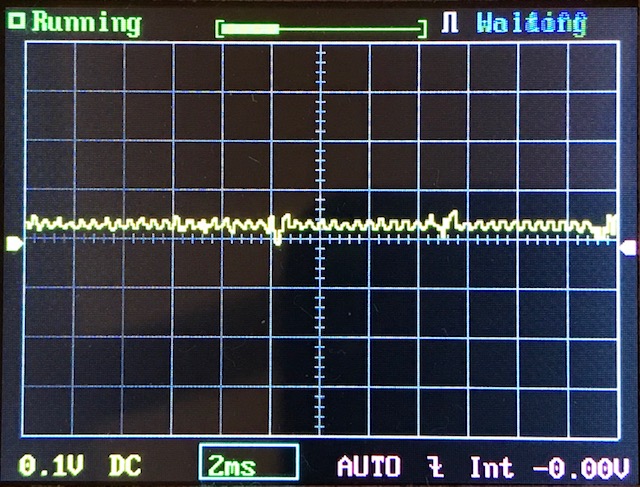 A digital oscilloscope screen displaying a mostly flat yellow waveform with minor fluctuations, grid lines, and status indicators. Text at the bottom shows settings: 0.1V, 2ms, DC, AUTO, and 0.00V.