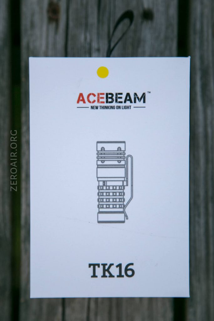 A white box labeled ACEBEAM with the slogan New Thinking On Light shows a line drawing of a flashlight and the model name TK16 beneath it. A yellow sticker with a black string is attached at the top.