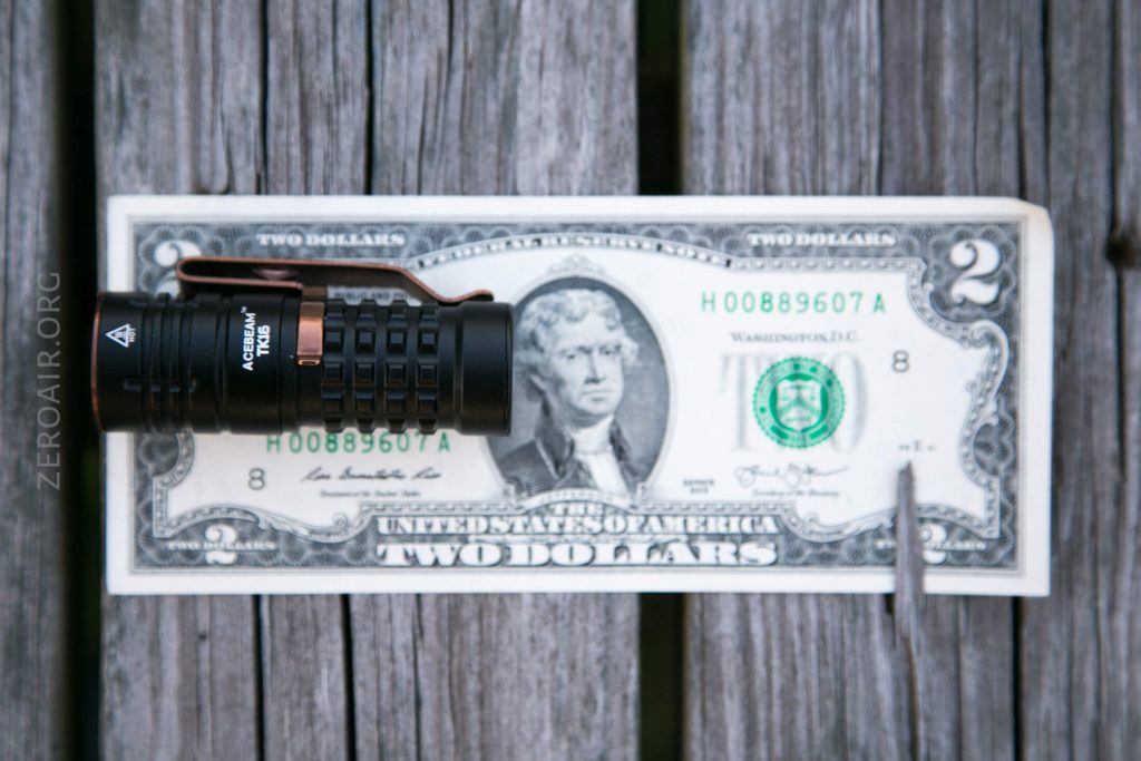 A small black flashlight and a silver pen are placed on top of a two dollar bill, which lies on a weathered wooden surface.