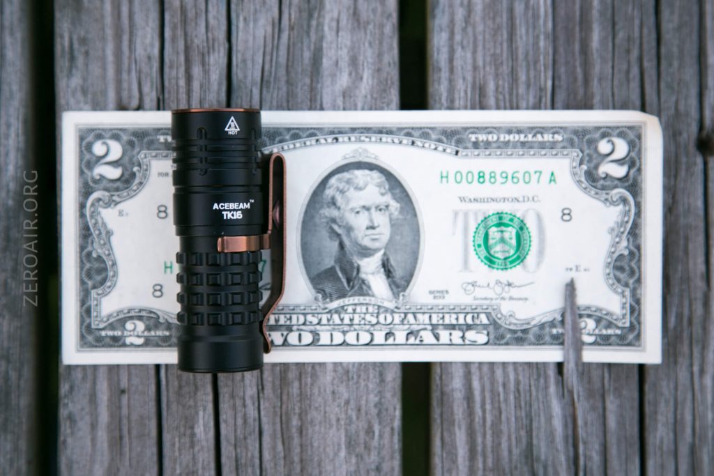 A black Acebeam TK16 flashlight is placed upright on a U.S. two-dollar bill, which rests on a weathered wooden surface. The flashlight covers part of Thomas Jefferson’s portrait on the bill.