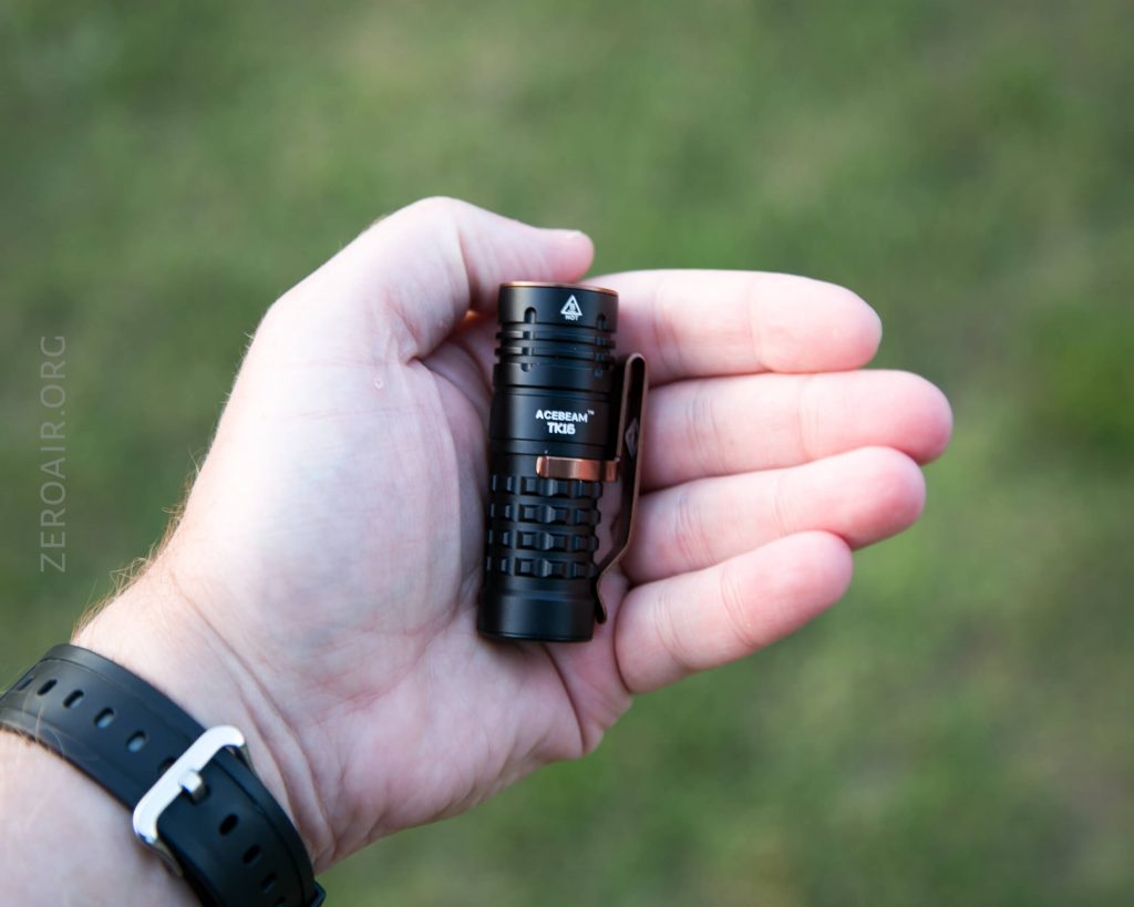 A person’s left hand holds a small black flashlight labeled “ACEBEAM TK16” against a blurred grassy background. The person wears a black watch with a white buckle on their wrist.