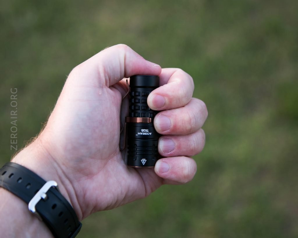 A person wearing a black wristwatch holds a small black flashlight in their hand against a blurred grassy background. The flashlight is labeled Wuben TO46R.