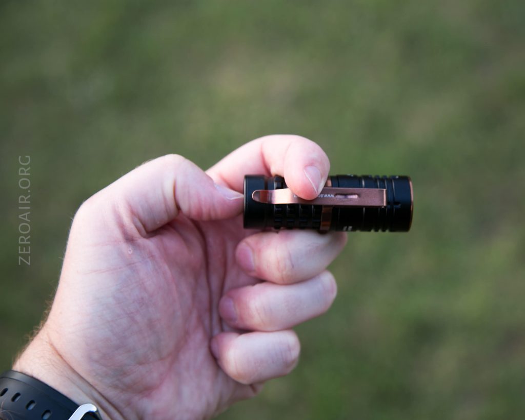 A hand holding a small black flashlight with a copper-colored clip, positioned against a blurred green outdoor background.