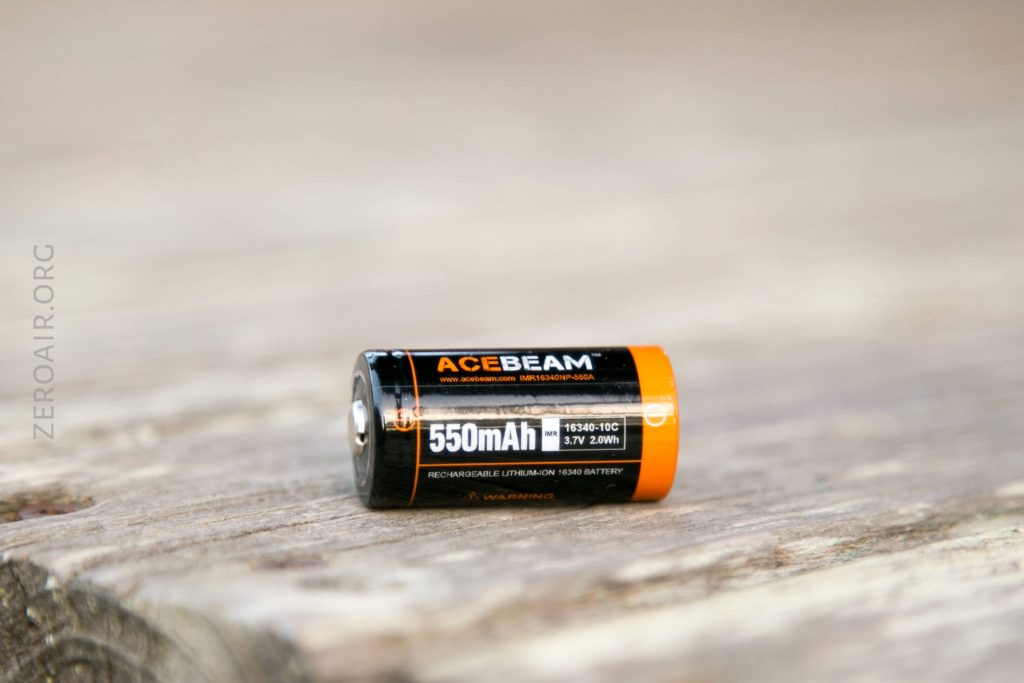 A close-up of an ACEBEAM 550mAh rechargeable lithium-ion battery (16340-10C, 3.7V 2.0Wh) resting on a weathered wooden surface. The battery label details are clearly visible.