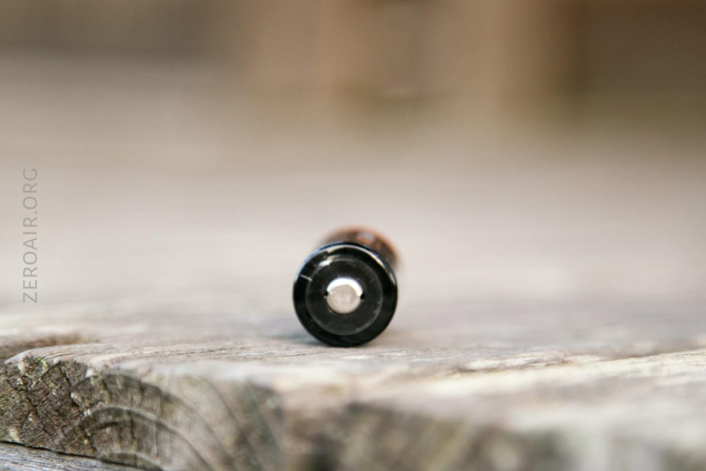 A single cylindrical battery lies horizontally on a wooden surface, with the positive terminal facing the camera. The background is blurred, and there is text reading ZEROAIR.ORG along the left side.