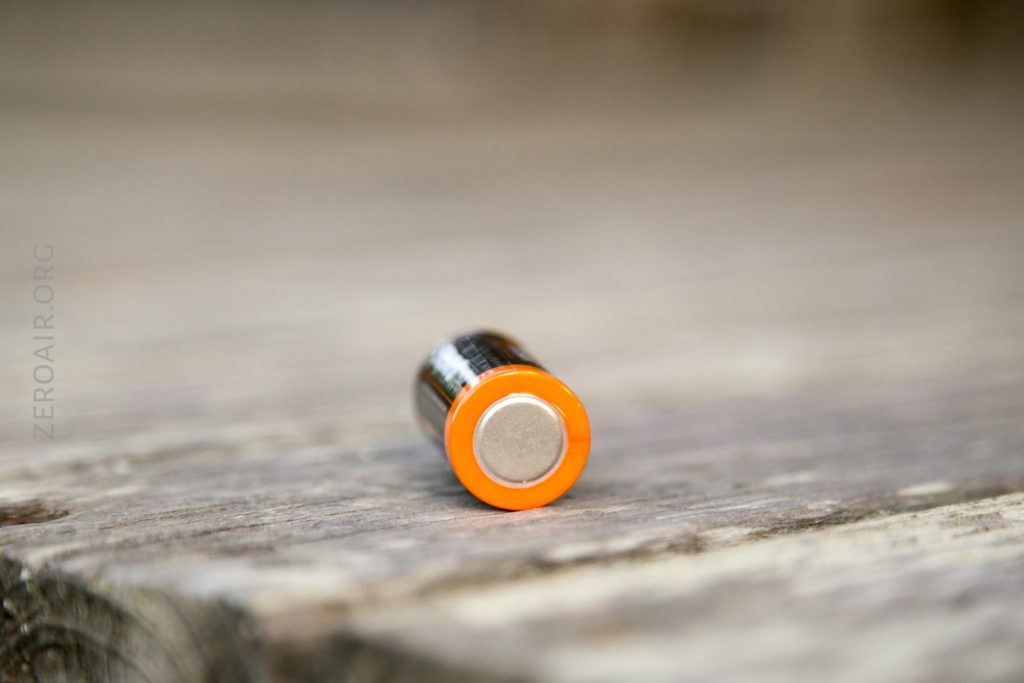 A cylindrical battery with an orange rim around the positive terminal is lying on its side on a wooden surface. The background is out of focus.