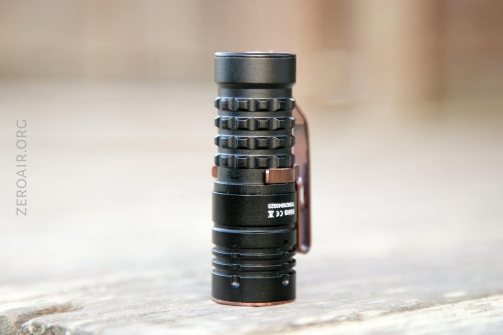 A small, black, cylindrical flashlight with a knurled grip pattern and a metal pocket clip stands upright on a wooden surface. The flashlight has white text and symbols on its side.