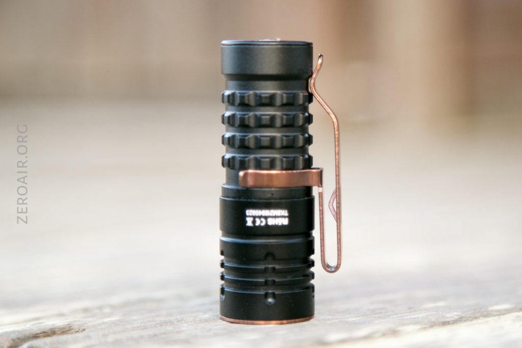 A small black flashlight with a textured grip and a metal clip stands upright on a light wooden surface. The flashlight is labeled EDC X and has ZEROAIR.ORG text faintly visible in the background.