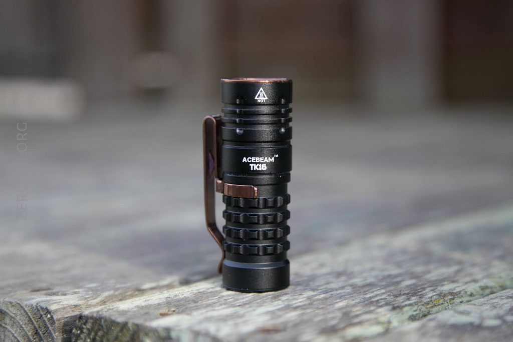 A compact black Acebeam TK16 flashlight stands upright on a weathered wooden surface with a blurred background.
