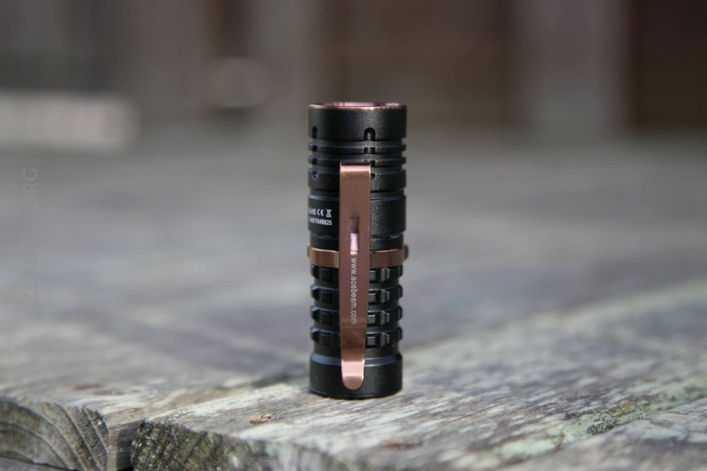 A black and bronze flashlight with a metal pocket clip stands upright on a weathered wooden surface, with a blurred background.
