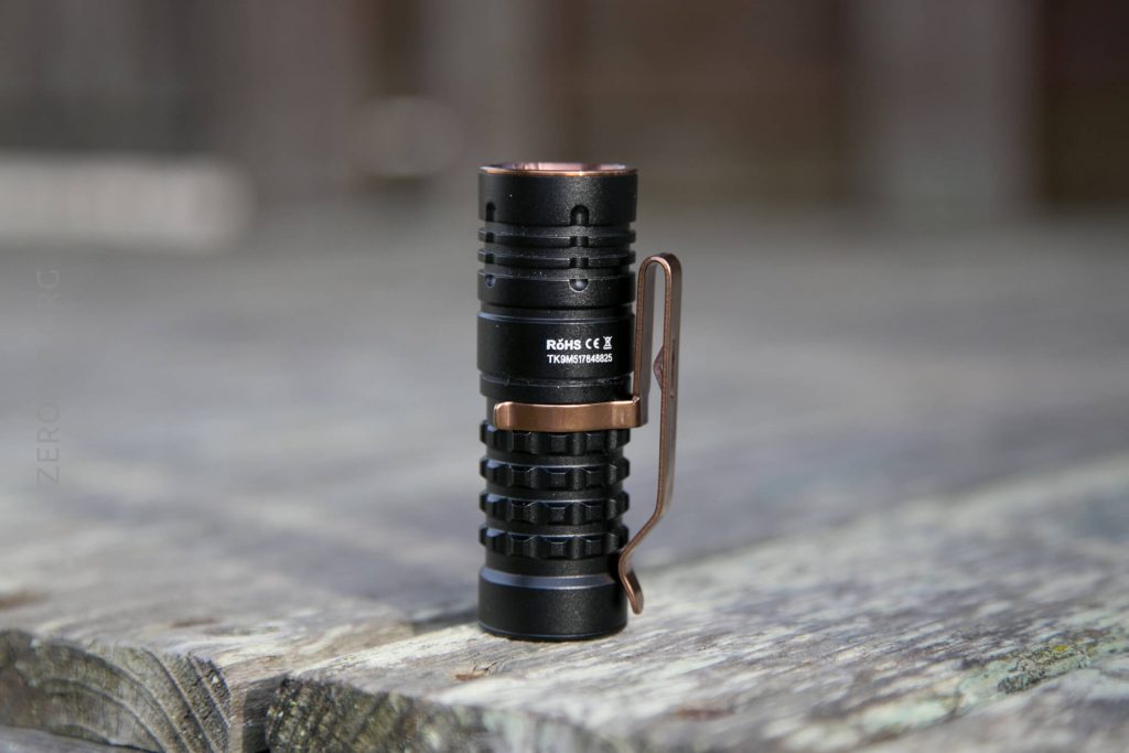 A small black flashlight with a bronze-colored clip and detailing stands upright on a weathered wooden surface outdoors.