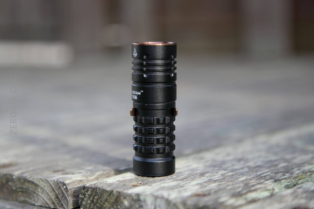 A black tactical flashlight with a textured grip and a copper-colored accent is standing upright on a weathered wooden surface. The background is blurred.