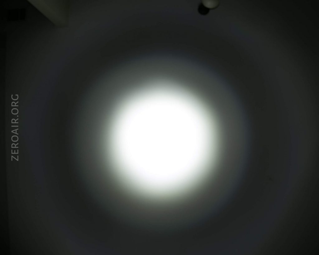 A bright white circular light beam shines on a dark surface, creating concentric rings of light and shadow. ZEROAIR.ORG is faintly visible on the left side of the image.
