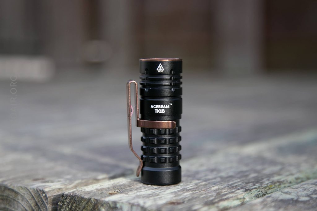 A compact black Acebeam TK16 flashlight with a side clip stands upright on a weathered wooden surface with a blurred background.