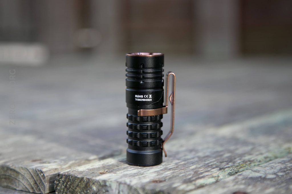 A compact black flashlight with a bronze clip stands upright on a weathered wooden surface, with a blurred background. The label on the flashlight reads Rohs CE X Tracpenbar.