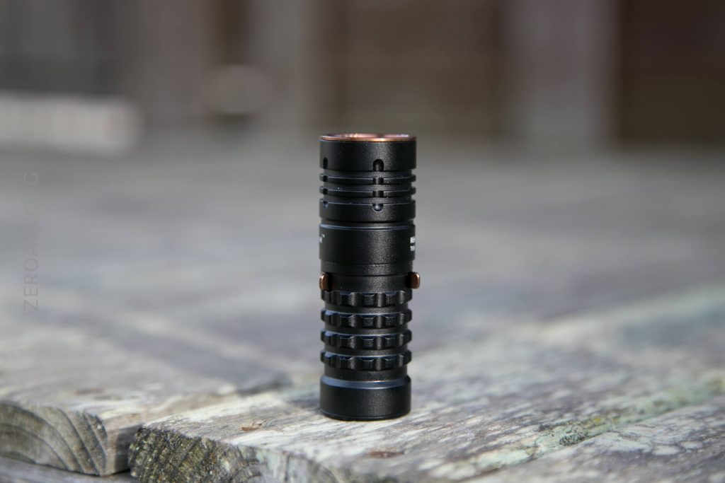 A small, black, cylindrical flashlight stands upright on a weathered wooden surface, with a blurred background.