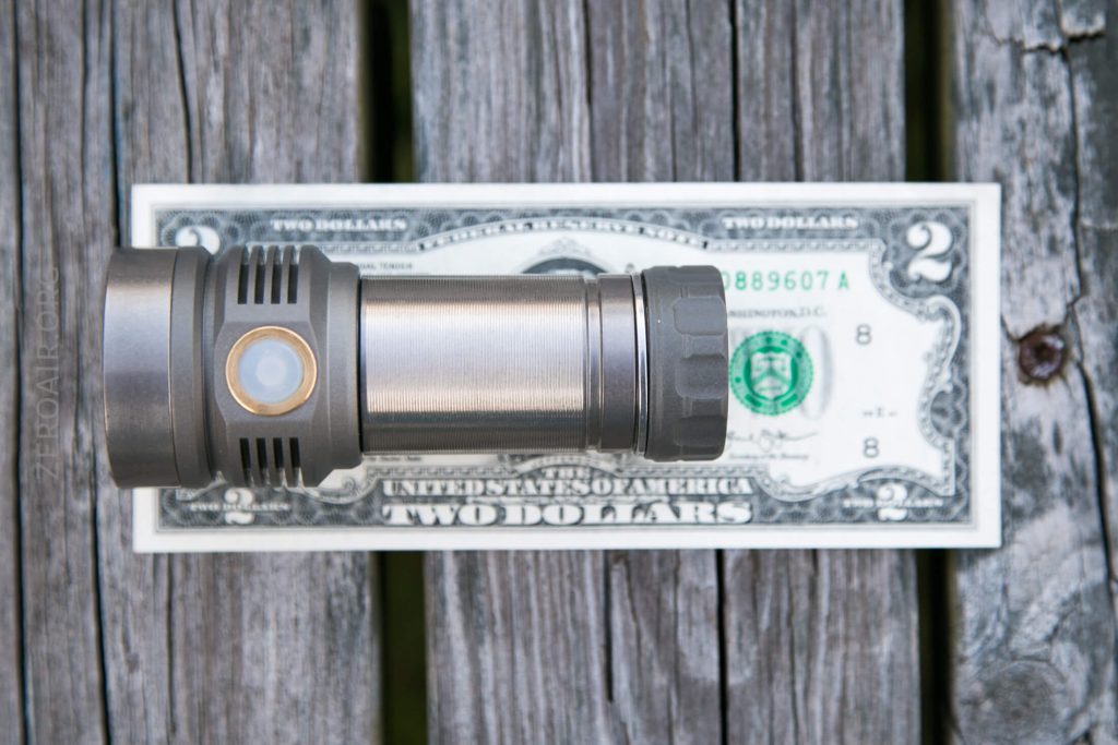 A metal flashlight is placed horizontally on top of a two-dollar bill, both resting on weathered wooden planks.