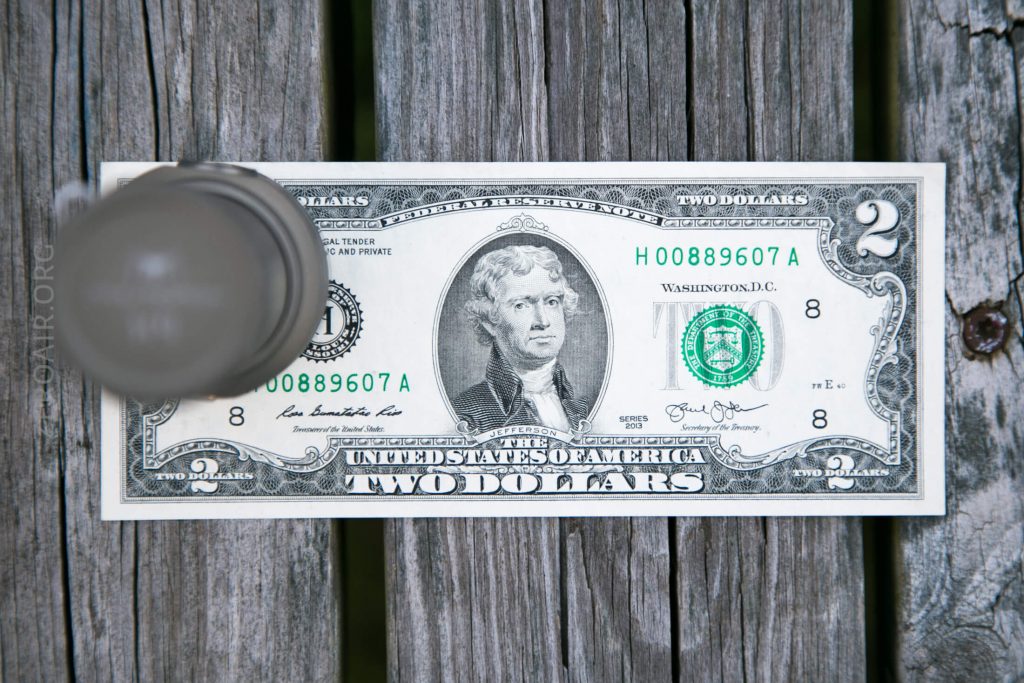 A two-dollar bill placed on a wooden surface with a round, semi-transparent object partially obscuring the left side of the bill.