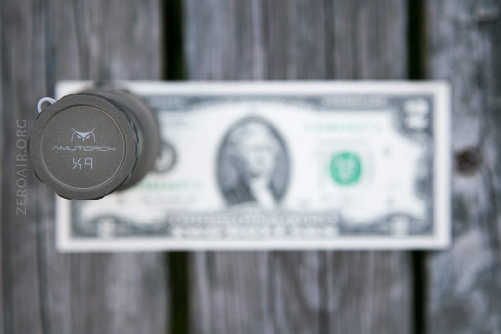 A top-down view of a flashlight labeled AMUTORCH X9 placed vertically on a US one-dollar bill, both set on a weathered wooden surface.