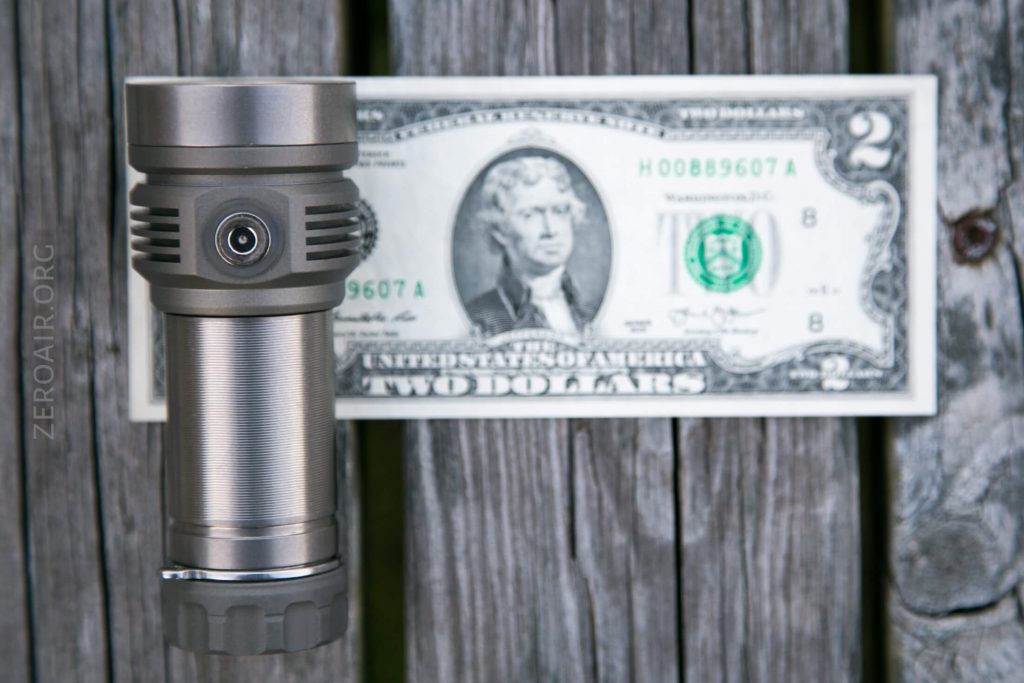 A metallic flashlight is placed vertically in front of a U.S. two-dollar bill, both set against a wooden plank background. The flashlight partially covers the left side of the bill.