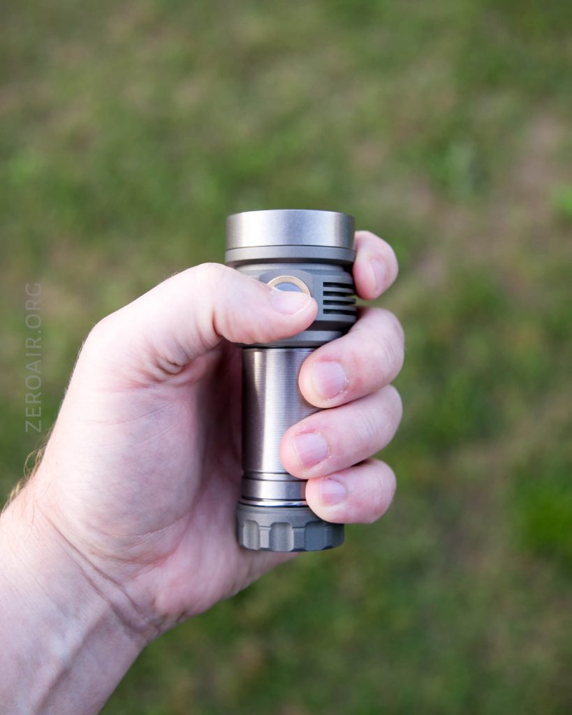 A person holding a metallic flashlight in their right hand against a blurred grassy background.