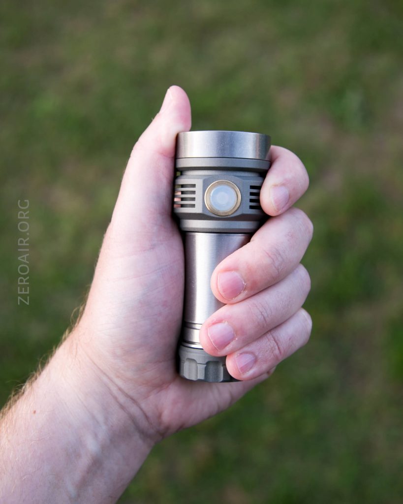 A left hand holding a metallic flashlight with a side button, shown against a blurred grass background. The flashlight is cylindrical and has grooves on its body. The watermark ZEROAIR.ORG is visible on the left side of the image.