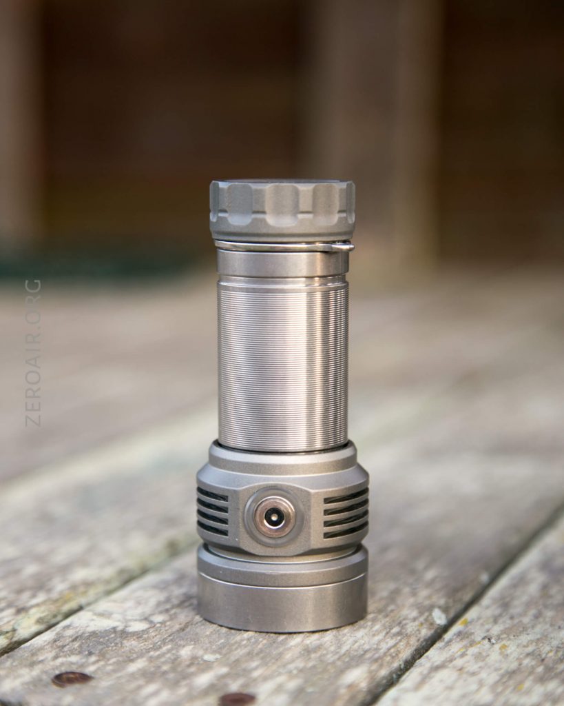 A metallic cylindrical flashlight with a textured grip and grooved head is standing upright on a weathered wooden surface. The background is out of focus.