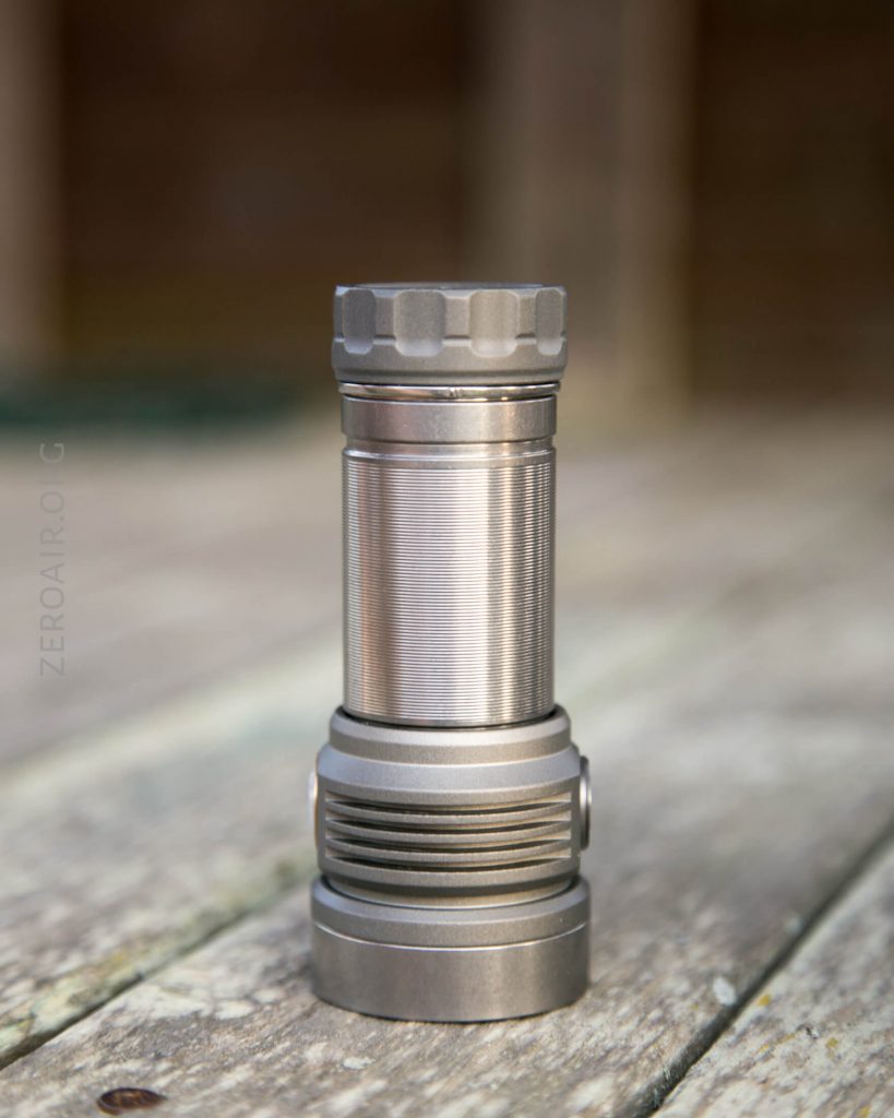A metallic flashlight is standing upright on a weathered wooden surface with a blurred brown and green background.