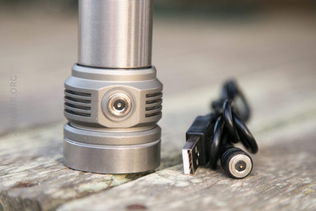 A close-up of the base of a metallic flashlight with a charging port, next to a coiled USB charging cable, both placed on a wooden surface.