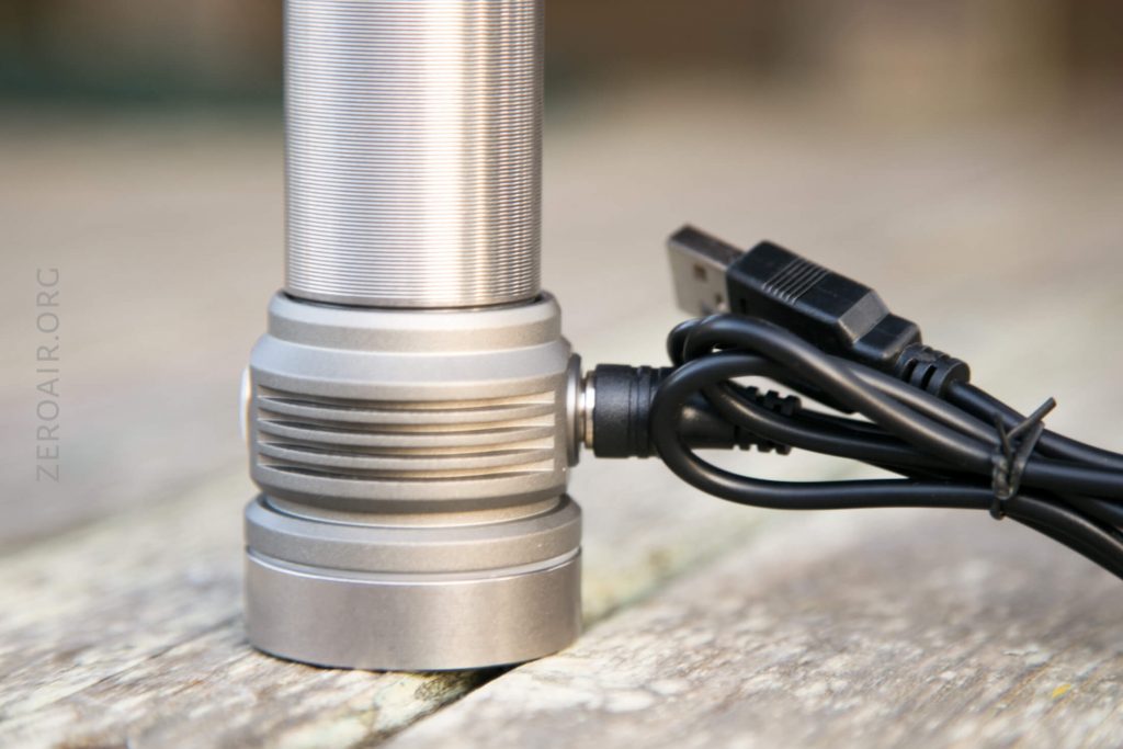 A close-up of a metallic flashlight standing upright on a wooden surface, with a USB charging cable plugged into its side.