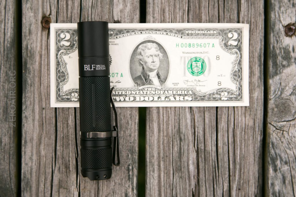 A black BLF Special Edition flashlight is placed on top of a U.S. two-dollar bill, both resting on a weathered wooden surface.