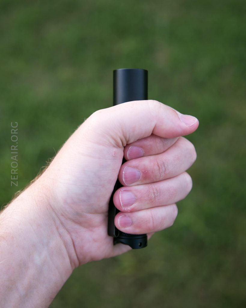 A close-up of a persons left hand holding a small black cylindrical object, possibly a flashlight, against a blurred grassy background. The website ZEROAIR.ORG is faintly visible on the left side.