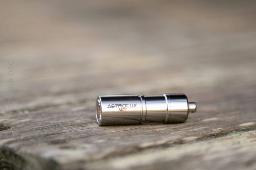 A small, metallic cylindrical flashlight labeled “ASTROLUX M01” lies on a weathered wooden surface. The flashlight is silver with a smooth finish and a slightly raised button on one end.