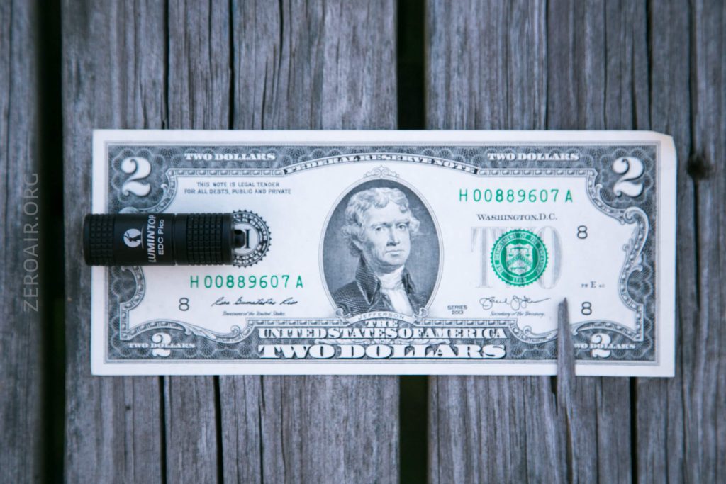 A two-dollar bill is placed on a weathered wooden surface with a small flashlight on the left side and a metal pen on the right side of the bill.