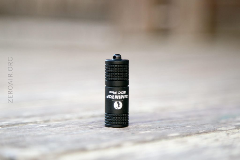 A small black cylindrical flashlight labeled LUMINTOP EDC Pico stands upright on a wooden surface. The background is out of focus. The text ZEROAIR.ORG is faintly visible on the left side of the image.