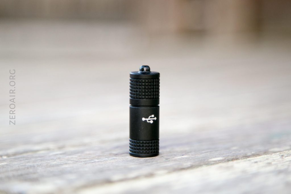 A small, black cylindrical device with textured ends and a USB symbol printed on the side, standing upright on a wooden surface. The background is out of focus, and ZEROAIR.ORG is visible on the left.
