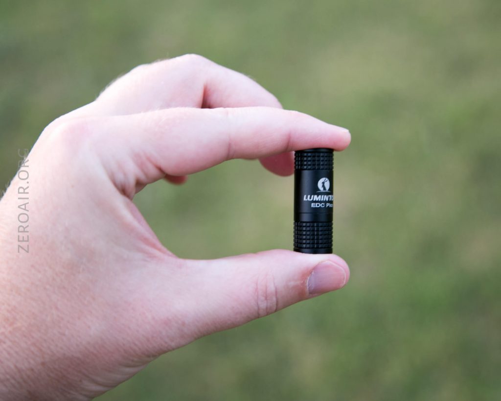 A person holds a small black cylindrical flashlight labeled LUMINTOP EDC Pico between their thumb and index finger, with a blurred grassy background.