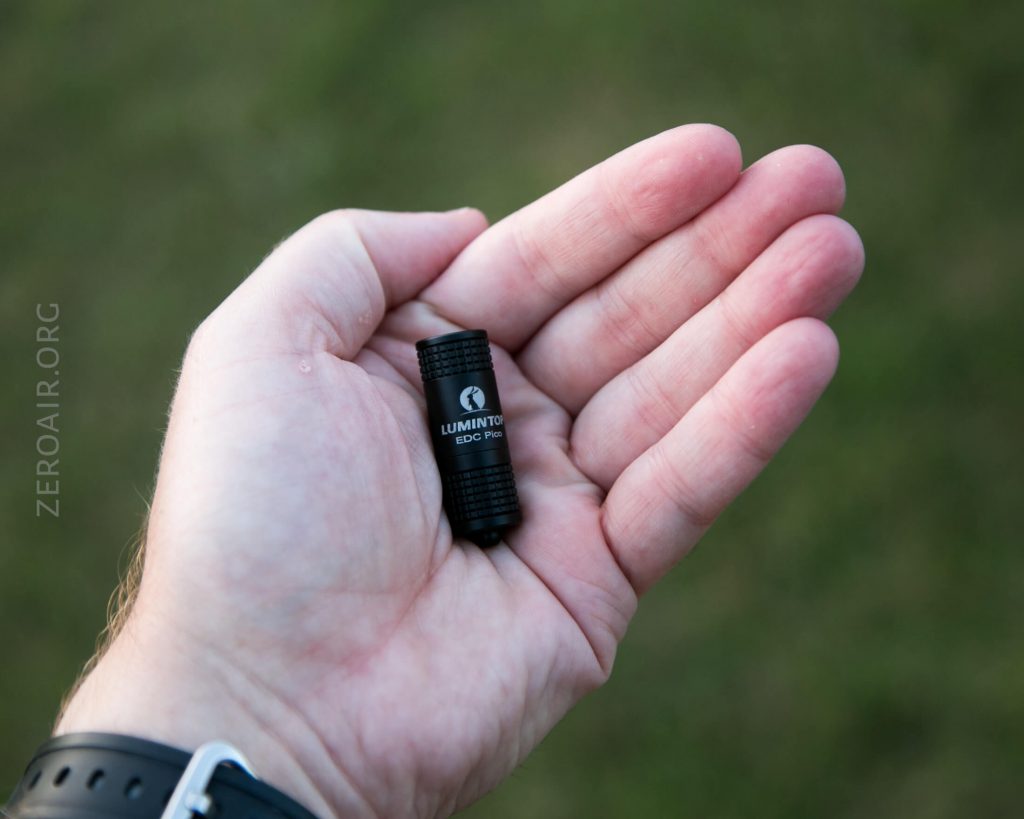 A person holds a small black LUMINTOP EDC Pico flashlight in their open palm against a blurred grassy background. The flashlight features ridged edges and is compact, fitting easily in the hand.