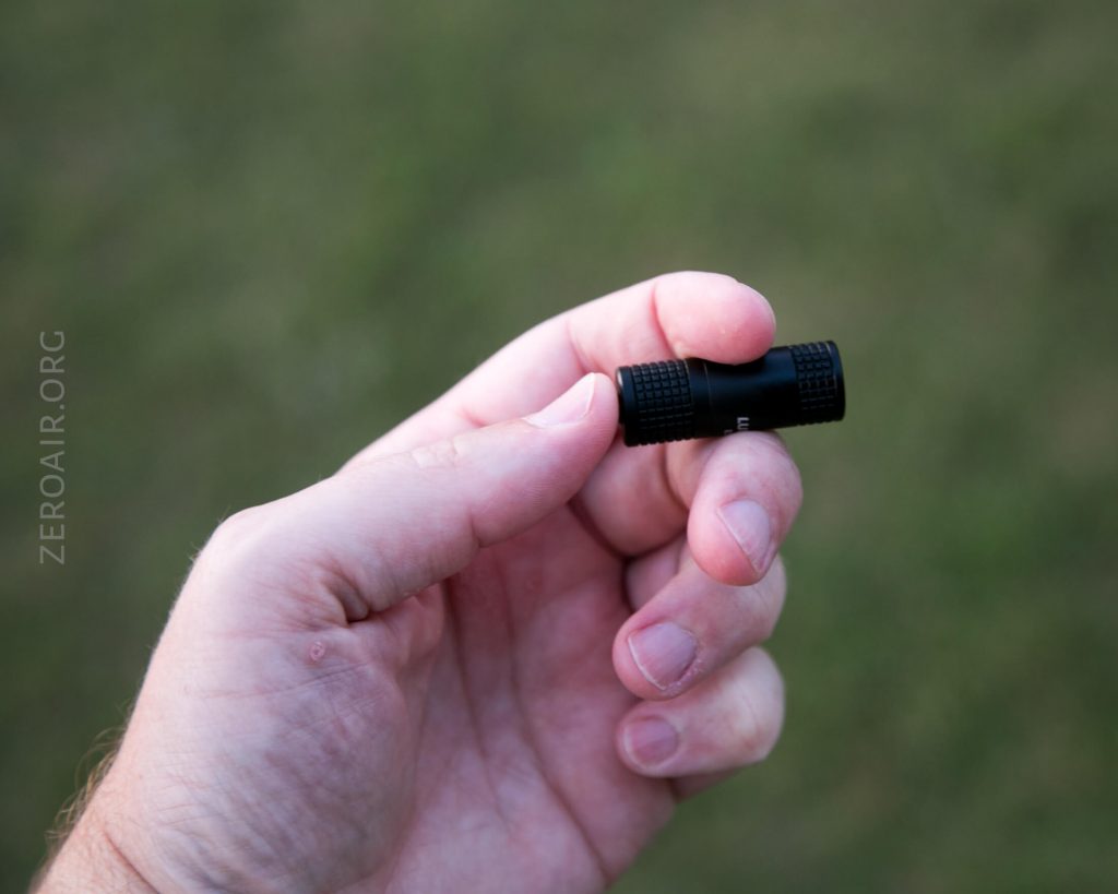 A hand is holding a small black cylindrical object, possibly a flashlight or tool, between the thumb and index finger against a blurred green background.